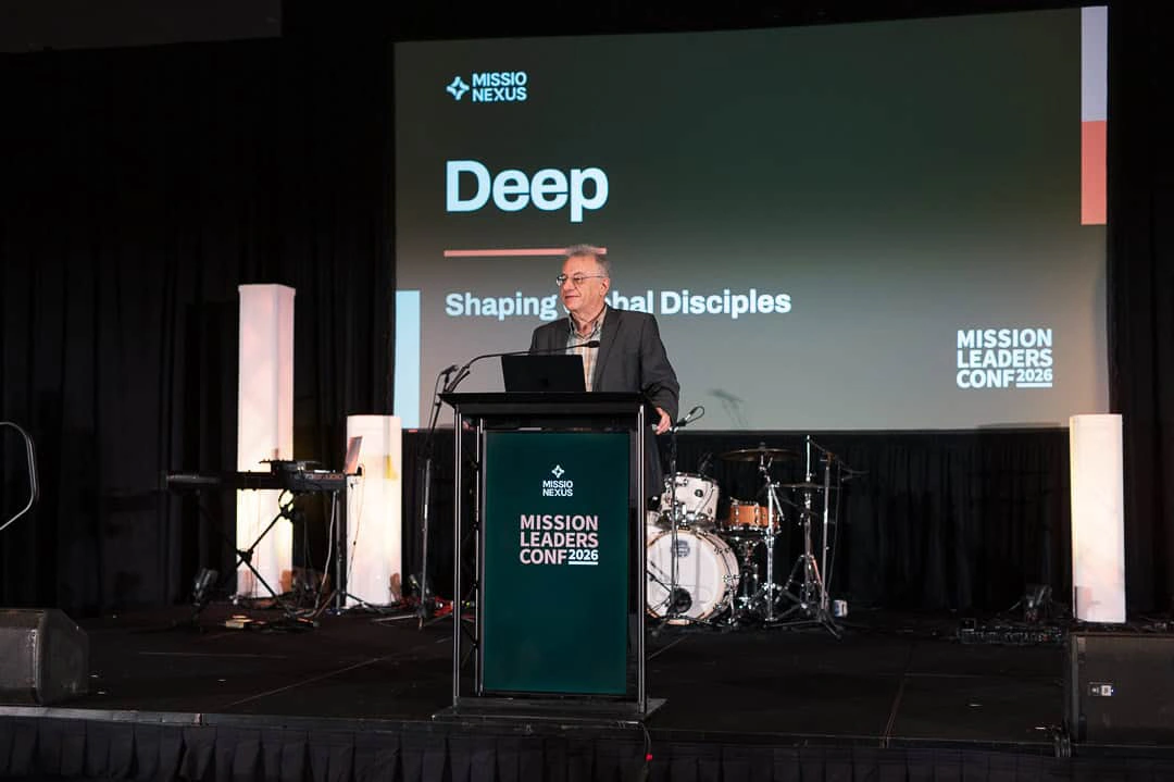A speaker at the podium in front of the Shaping Global Disciples slide
