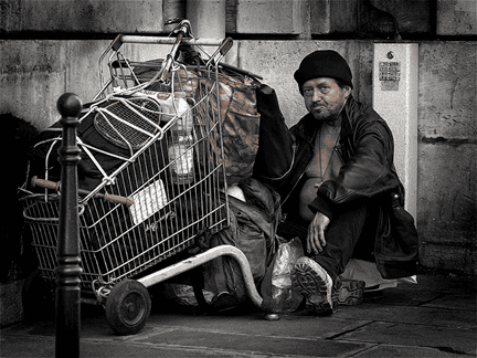 Homeless man resting beside shopping cart filled with belongings on city street.