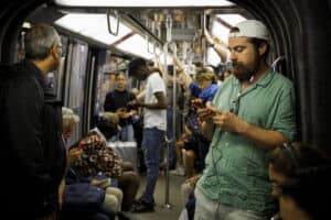 Subway passengers on a train, some using smartphones, in a busy urban transit setting.