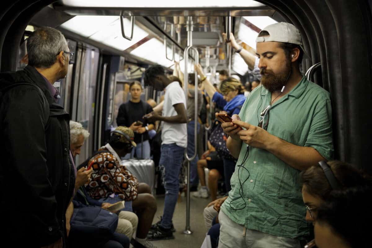 Subway passengers on a train, some using smartphones, in a busy urban transit setting.