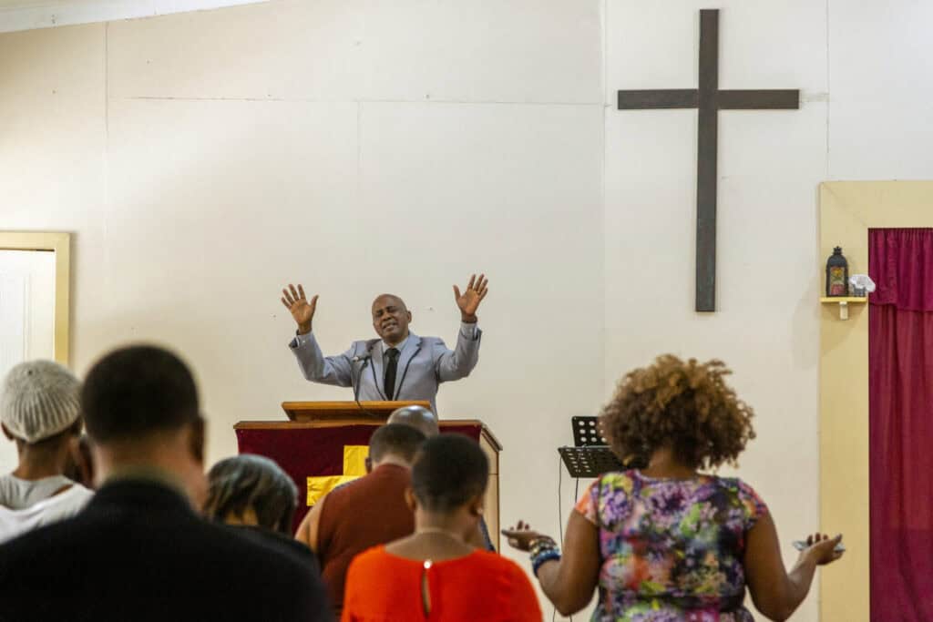 Pastor speaking passionately to congregation in church setting with cross in background.