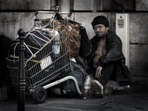 Homeless man resting beside shopping cart filled with belongings on city street.