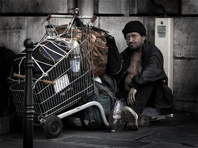 Homeless man resting beside shopping cart filled with belongings on city street.