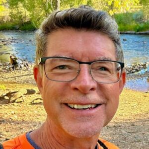 Smiling man with glasses outdoors near a river in a natural setting.
