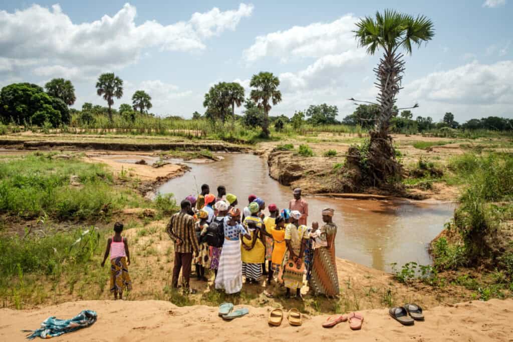 Community gathering by a river in a rural landscape with palm trees.
