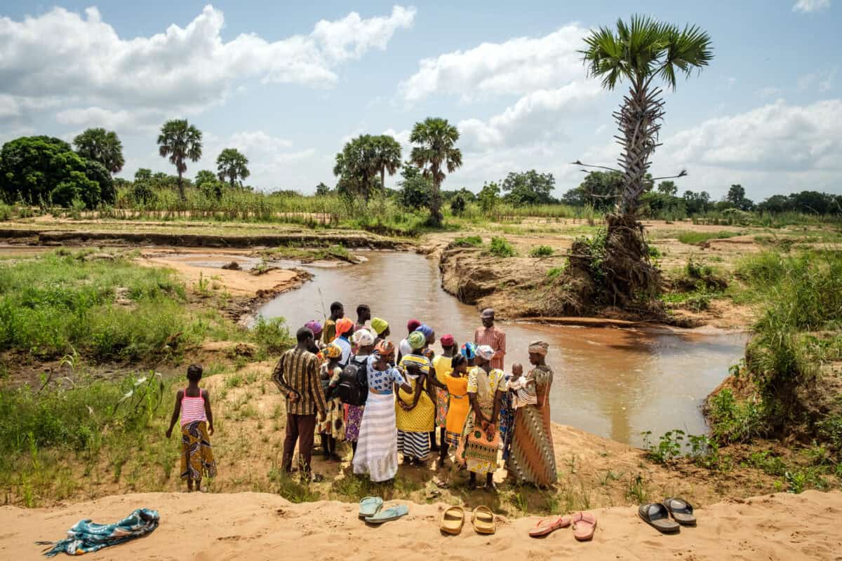Community gathering by a river in a rural landscape with palm trees.