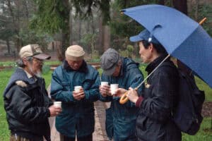 Group of people sharing drinks outdoors in the rain with umbrellas and raincoats.